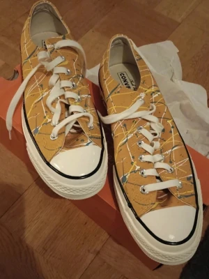 Gula Converse All Star Chuck Taylor sneakers Storlek 44,5 - Unika Converse All Star sneakers i gult med färgstänk och vita detaljer. Klassisk låg modell med vit tå och snörning. Snyggt mönster med stänk i blått, vitt och gult på canvasmaterial. Perfekta för dig som vill sticka ut.