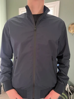 Mörkblå bomberjacka från J.Lindeberg - Snygg mörkblå vind/bomberjacka från J.Lindeberg med minimalistisk design. Jackan har hög krage, hel dragkedja framtill och två snedställda fickor med dragkedja. Mycket bra skick, utan defekter. Passar lika bra till golfbanan som till en vardaglig stilren ”look”. Köpt för 1600 då den köptes på rea👍skriv om du vill diskutera priset vid snabb affär 📫 