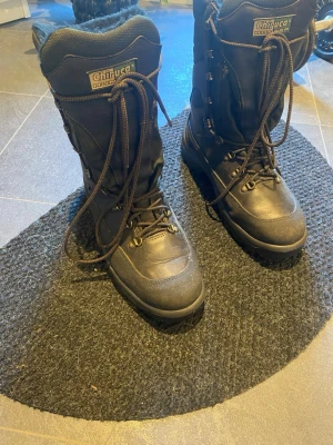 Chiruca svarta fodrade vinterkängor - Svarta vinterkängor från Chiruca med högt skaft, grov sula och snörning hela vägen upp. Kängorna har varm, mjuk fodring och slitstarkt yttertyg i en mix av läder och textil. Perfekta för kalla dagar och outdoor-äventyr. Märket syns tydligt på plösen och sidan.