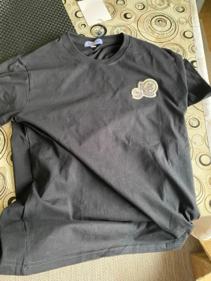 Svart Moncler t-shirt med patch - Snygg svart t-shirt från Moncler med rund hals och två broderade patches på bröstet i blått, beige och rött. Tillverkad i mjuk bomull och har en klassisk passform som funkar till det mesta. Perfekt för dig som gillar stilrena plagg med exklusiv känsla. Pris kan diskuteras vid snabb köp.