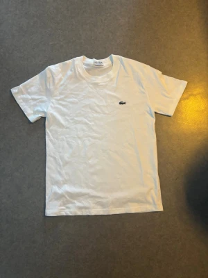 Vit t-shirt från Lacoste - Klassisk vit t-shirt från Lacoste med den ikoniska krokodilloggan broderad på bröstet. T-shirten har rund halsringning och korta ärmar. Tillverkad i mjuk bomull för en skön och avslappnad passform.