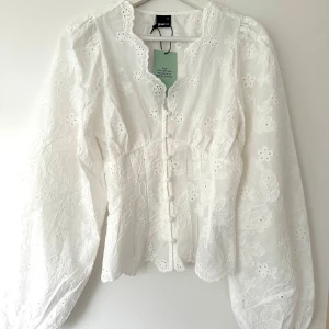 Vit broderad blus från gina tricot - Romantisk vit blus från gina med broderade blommor och dekorativa knappar framtill. Blusen har långärmade puffärmar, v-ringad hals med vågig kant och är tillverkad i ett lätt, luftigt tyg. Perfekt för en trendig och feminin look.