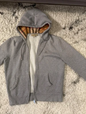 Burberry Zip Hoodie - Säljer en grå hoodie från Burberry med klassiskt rutigt foder i huvan och mjuk vit fleece på insidan. Hoodien har dragkedja, två fickor fram och justerbar huva med snören. Perfekt för dig som gillar stilrena och exklusiva plagg. Självklart äkta. 