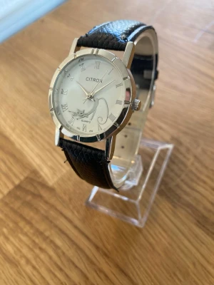Citron ”The Cat Lady” 32mm Damur - Elegant mycket ovanlig variant av Citron S565S Quartz armbandsur som har ett klassiskt utseende. Klocka är i nyskick enligt bilder och har en silverfärgad boett och en vit urtavla med romerska siffror. Visarna är silverfärgade och en stretchande katt också i silver. Armbandet är tillverkat av svart läder med en texturerad yta och har en silverfärgad spänne. Baksidan av boetten är i rostfritt stål. Behöver bara nytt batteri.  Nypris: 500 SEK Mitt pris: 189 SEK  Skickas alltid snabbt och spårbart.