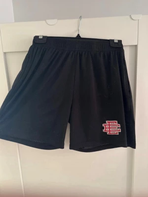 EE Shorts  ( Eric Emanuel) Äkta - Säljer ett par svarta meshshorts med elastisk midja och broderad rödvit logga på vänster ben. Shortsen har en luftig passform och är perfekta för sport eller chill. Materialet är lätt och andas, vilket gör dem sköna under varma dagar.