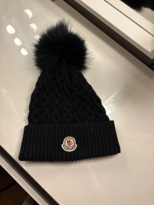 Svart Moncler mössa med pälsboll - Snygg svart stickad mössa från Moncler med stor fluffig pälsboll på toppen och klassisk Moncler-logga framtill. Mössan har ribbad kant och kabelstickat mönster som ger en trendig look. Perfekt accessoar för kalla dagar.