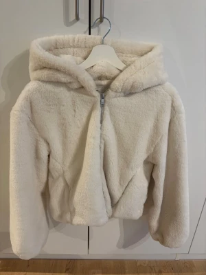 Vit teddyjacka med huva - Supermjuk och fluffig teddyjacka i vit färg med dragkedja framtill och stor huva. Jackan har en avslappnad passform och långa ärmar, perfekt för kalla dagar. Tillverkad i fuskpäls som ger en cozy vibe.