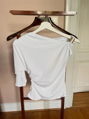 Vit oneshoulder topp från Zara - Säljer en vit oneshoulder topp från Zara med snygg drapering och lätt rynkning i sidan. Toppen har trekvartsärm på ena sidan och är axelbandslös på andra. Helt oanvänd då den inte passade och jag inte kunde skicka tillbaka den💗💗