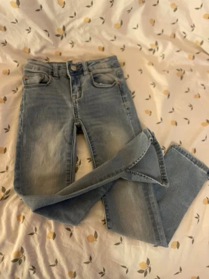 Ljusblå raka jeans barn st 130 - Säljer ett par ljusblå jeans med raka ben och klassisk femficksmodell. Jeansen har en snygg tvätt med slitningar framtill och stängs med knapp och dragkedja. Barn storlek 130