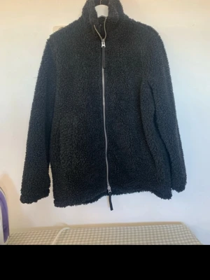 Gråmelerad  teddyjacka med dragkedja - Mysig gråmelerad  teddyjacka med hög krage och hel dragkedja framtill. Jackan har två sidofickor och är gjord i fluffigt, mjukt material som håller dig varm. Perfekt för kyliga dagar och ger en avslappnad, trendig look. Nypris 1199:-