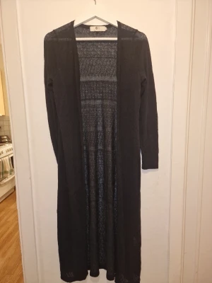 Svart lång kofta - Svart lång tunnstickad kofta med diskret mönster. Snygg att slänga över en outfit för extra lager. Enkel och stilren design som passar till mycket.
