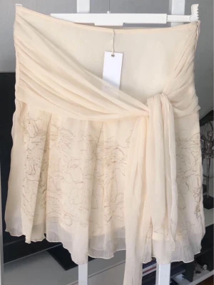 Beige fejk omlottkjol med volang NY med prislappen  - Säljer en ljusbeige omlottkjol med volangdetaljer och knytband i midjan. Kjolen har ett skirt, lätt material och ett subtilt blommigt mönster framtill. Perfekt för sommarens alla tillfällen! Midjemått ca 78, dragkedja på sidan 