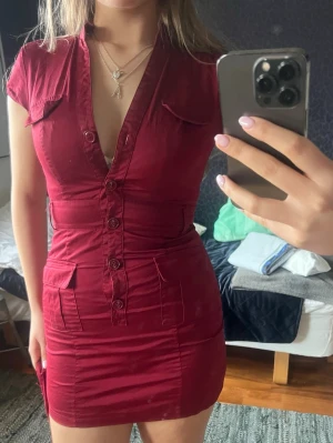 Vinröd kort klänning från H&M - Kort vinröd klänning från H&M med knappar framtill, två bröstfickor med lock och tillhörande bälte i midjan. Klänningen har korta ärmar och ståkrage, tillverkad i bomull för en bekväm känsla.