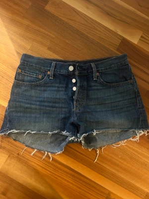 Levi's blå jeansshorts med rå kant - Säljer ett par klassiska blå jeansshorts från Levi's med hög midja och rå, fransig kant. Shortsen har fem fickor, knappar framtill och den ikoniska Levi's-lappen bak. Perfekta för sommaren och ger en avslappnad vibe. Uppskattar till XS? vet ej. se bilder.