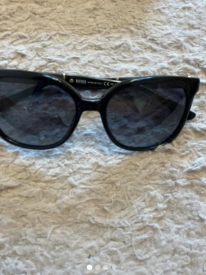 Svarta solglasögon med gulddetaljer från Hugo Boss - Snygga solglasögon i svart med klassisk rektangulär form och tunna guldfärgade detaljer vid bågarna. Perfekta för att höja din stil på sommaren. Passar till de flesta outfits och ger en cool vibe.