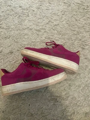 Vinröda Nike Air Force 1 sneakers - Säljer ett par vinröda Nike Air Force 1 sneakers i mocka med beige detaljer på hälen och vit sula. Klassisk låg modell med snörning och tydlig swoosh på sidan. Perfekta för dig som vill sticka ut med färg på fötterna.