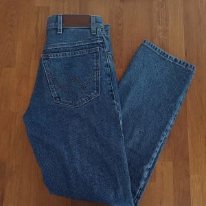 Blå jeans från Wrangler - Klassiska blå jeans från Wrangler med rak passform och hög midja. Jeansen har den ikoniska W-sömmen på bakfickan och är gjorda i slitstark denim. Perfekta för en avslappnad och tidlös stil.