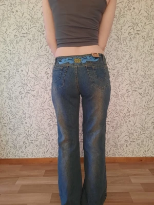 Blå Zodiac 2003 bootcut jeans - Säljer ett par blå bootcut jeans från Zodiac med broderade detaljer i ljusblått och en unik patch på ena benet. Jeansen har klassisk femficksmodell, låg midja och en cool retrovibe. Materialet är denim i bomull och passformen är avslappnad.