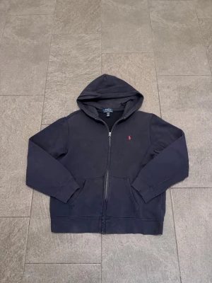 Mörkblå hoodie från Polo Ralph Lauren - Mörkblå hoodie med dragkedja från Polo Ralph Lauren. Klassisk design med huva, två fickor framtill och den ikoniska röda logotypen broderad på bröstet. Tillverkad i mjukt bomullsmaterial som är skönt att bära. Perfekt för en avslappnad och stilren look. Med ett litet hål i armen. Den är XL i barn storlek men i vuxen är den typ S-M