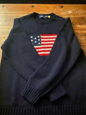 Mörkblå stickad tröja Polo Ralph Lauren - Mörkblå stickad tröja från Polo Ralph Lauren med amerikansk flagga på bröstet. Tröjan har rund halsringning, ribbstickade muddar och nederkant. 