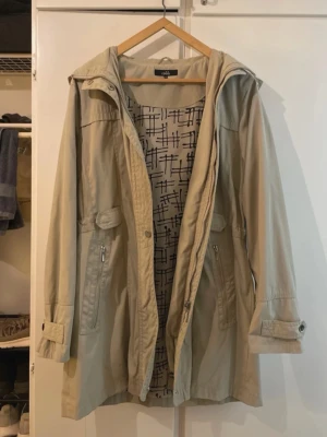Beige/khaki Isolde Parkasjacka - Stilren beige parkasjacka från Isolde med justerbar midja, dragkedja och knappar framtill. Jackan har två fickor med dragkedja, dekorativa knappar vid ärmslut och en praktisk huva. Fodret har ett grafiskt svartvitt mönster som ger extra karaktär. Använd några gånger och är i bra skick.   Som ses på sista bilden har ena huvknappen gått loss.
