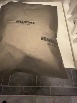 Grå hoodie från Essentials Fear of God - Säljer en stilren grå hoodie från Essentials Fear of God. Hoodien har en klassisk passform med huva och är tillverkad i mjukt bomullsmaterial. Diskret logga på bröstet och en svart patch med märkets namn på ryggen. Perfekt för en clean och avslappnad look.