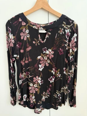 KAFFE - Blommig långärmad blus i svart - Snygg svart blus med blommigt mönster i rosa, vitt och brunt. Blusen har långa ärmar och en liten v-ringning framtill. Materialet känns mjukt och lätt, perfekt för en trendig vardagslook. Passar till både jeans och kjol.