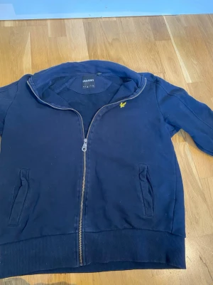 Marinblå hoodie från Lyle & Scott - Snygg marinblå hoodie från Lyle & Scott med dragkedja framtill och två fickor. Klassisk gul logga på bröstet och ribbade muddar vid ärmslut och nederkant. Perfekt för en avslappnad stil och enkel att matcha med jeans eller joggers.