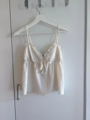 Linnetopp, Nelly  - Säljer en söt beige/krämvit linnetopp från Nelly. Toppen har tunna justerbara axelband och en v-ringning med volangkant och en knytdetalj framtill. Toppen är tillverkad i ett lätt, strukturerat och luftigt material som passar perfekt till sommaren. (Använd endast ett fåtal gånger)