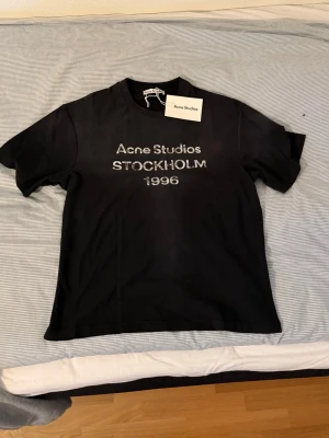 Black Acne Studios t-shirt - Snygg svart t-shirt från Acne Studios med tryck 'Acne Studios STOCKHOLM 1996' på bröstet. Klassisk passform med rund hals och korta ärmar. Tillverkad i mjuk bomull och perfekt för en clean och stilren look. Helt ny storlek S