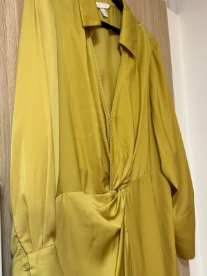 Gul omlottklänning från H&M - Säljer en snygg gul omlottklänning från H&M med lång ärm och krage. Klänningen har en twistad detalj i midjan och är gjord i ett glansigt, mjukt material som ger en lyxig känsla. Perfekt för dig som vill sticka ut med färg och stil.