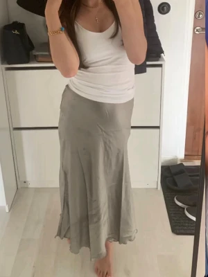 Grå långkjol i satin, storlek XS - Säljer en snygg grå långkjol i satin. Kjolen har en mjuk, glansig yta och faller fint längs benen. Perfekt för dig som gillar en enkel men stilren look. Passar till både sneakers och klackar.