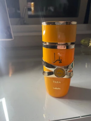 Lattafa Yara parfym orange flaska - Lattafa Yara parfym i en snygg orange flaska med silverdetaljer och arabiska bokstäver. Flaskan har en unik form med dekorativa band och ett emblem i mitten. Perfekt för dig som gillar exklusiva och trendiga dofter. 95% kvar