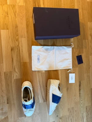 Valentino vita och blå sneakers - Snygga vita sneakers från Valentino med bred blå rem över ovandelen. Skorna har klassisk låg siluett, vita snören och detaljer i läder. Kommer med originalkartong, dustbag och certifikat. Perfekta för dig som vill ha en exklusiv touch på din outfit.