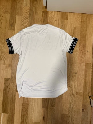 Vit Ellesse t-shirt med logga - Vit t-shirt från Ellesse med stor logga i svart, orange och rött på bröstet. Ärmarna har svarta muddar med Ellesse-tryck i vitt. Klassisk rak passform och rund hals. Tillverkad i mjukt och stretchigt material, perfekt för sport. Sitter tight runt armarna.