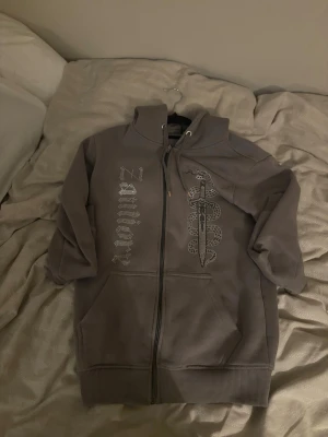 Zantiore zipper hoodie - Säljer en grå zip-hoodie med tryck av en orm som slingrar sig runt ett svärd på framsidan och texten 'Zillionz' i silvrigt tryck. Hoodien har fickor fram, ribbade muddar och en stor huva. Perfekt för dig som gillar streetwear och unika prints.