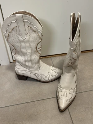 Vita cowboy boots med broderi - Säljer ett par vita cowboy boots i läder med snygga beige broderier och spetsig tå. De har klassisk hög skaftdesign, brunt klackparti och dekorativa sömmar längs hela skon. Perfekta för dig som vill sticka ut med westernstil.