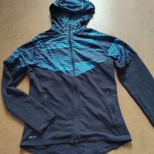 Nike Vindjacka Mörkblå - En mörkblå/blå vindjacka med luva från Nike. Hög krage, luva, dragsko runt luvan och bak på luvan. Reflex på ryggen. Perfekt för träning eller till vardags. Bra dragkedja och fint skick! Storlek M för damer men något liten i storleken. Dri-fit teknologi. 