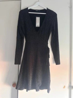 Svart glittrig omlottklänning Gina Tricot - Svart omlottklänning glittrigt tyg och knyt i midjan. Klänningen har v-ringning och långa ärmar
