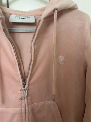 Juicy Couture - Snygg rosa hoodie i mjuk velour från Juicy Couture. Dragkedja med ikoniskt J-hänge, broderad logga på bröstet och fickor framtill. Klassisk huva och ribbade muddar. Perfekt för en chill och trendig look.