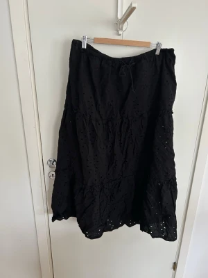 Svart långkjol med broderie anglaise - Svart kjol i midi med broderie anglais. Kjolen har resår och knytband i midjan för skön passform. Tillverkad i ett lätt bomullstyg som passar perfekt till varmare dagar. Stilren och enkel att matcha med olika toppar.