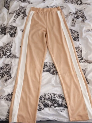 Beige mjukisbyxor med vita ränder - Snygga beige mjukisbyxor med raka ben och vita sidoremsor längs båda benen. Byxorna har resår i midjan för skön passform och är gjorda i ett mjukt, stretchigt material. Perfekta för en avslappnad och trendig look.