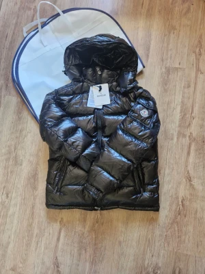 Svart Moncler Jacka - Snygg svart dunjacka från Moncler med glansig finish och stor huva. Jackan har två dragkedjefickor fram, Moncler-logga på ärmen och quiltad design. Perfekt för kalla vinterdagar och riktigt trendig look. Storlek 4.