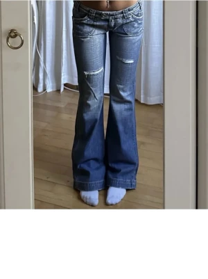 Blå bootcut jeans med slitningar - Säljer ett par blå bootcut jeans med slitningar på låret och snygga detaljer på fickorna. Jeansen har låg midja och klassisk femficksdesign. Perfekta för dig som gillar en avslappnad men trendig look. Innerben 80, midja 84❤️