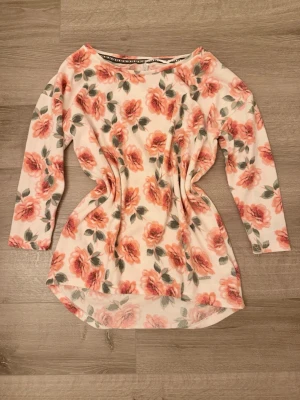 Blommig mjuk långärmad topp  - Supersöt långärmad topp från Hailys med blommigt mönster i rosa och grönt på vit botten. Toppen har rund halsringning och något längre bakstycke. Perfekt för dig som gillar romantisk och feminin stil.