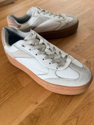 Vita sneakers med beige sula - Storlek 40 - Snygga vita sneakers med beige/brun platåsula och detaljer i mockaimitation. Skorna har klassisk låg design, vita skosnören och paneler i både läder och mocka-look. Perfekta för en clean och trendig stil.