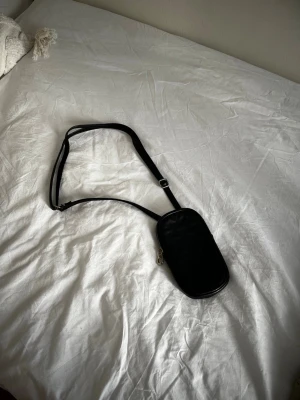 Svart axelväska med justerbart band - Snygg svart axelväska i minimalistisk design med justerbart axelband och silverfärgad dragkedja. Väskan har en avlång, rundad form och passar perfekt för det viktigaste. Materialet ser ut att vara konstläder.