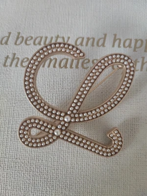 Brosch med pärldekor i guldton - Snygg brosch i form av en snirklig bokstav, dekorerad med små vita pärlor på en guldfärgad metallbas. Perfekt accessoar för att ge din outfit en personlig touch. Broschen har en säkerhetsnål på baksidan för enkel fastsättning.