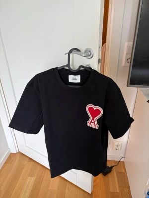 Svart Ami t-shirt med röd hjärtlogga - Snygg svart t-shirt från Ami med stor röd hjärtlogga på bröstet. Klassisk rak passform och rund halsringning. Tillverkad i mjukt bomullstyg som känns skönt mot huden. Perfekt för dig som gillar stilrena plagg med en twist.