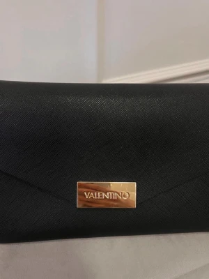Svart kuvertväska från Valentino - Stilren svart kuvertväska från Valentino med guldfärgad logga framtill. Väskan har en elegant, slät yta och magnetlås i guld. Insidan är enkel och rymlig, perfekt för det viktigaste. Kedja i guldton medföljer för att bära över axeln.